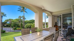 A covered patio with a table and chairs, overlooking a street and greenery. - Palms at Wailea #1605 Tropical Garden View Ground Floor, Wifi, Shared Hot Tub (Wailea)