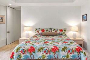 A bedroom with a bed covered in a floral bedspread, two bedside lamps, and a door to the left. - Palms at Wailea #1605 Tropical Garden View Ground Floor, Wifi, Shared Hot Tub (Wailea)