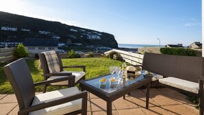 Une terrasse extérieure aménagée avec des meubles en osier, une table en verre et une vue sur la mer et les bâtiments qui se dressent à l’arrière-plan.