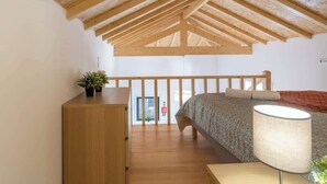 Een slaapkamer met een schuine kap, houten balken, een bed met beddengoed met een patroon, een nachtkastje en een lamp.