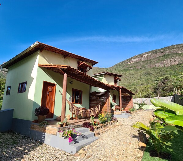 A two-story house with a wooden porch, a small garden with various plants, and a mountainous backdrop. - Green Chales (Palmeiras)