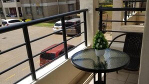 A balcony with a glass table and two chairs, overlooking a street with parked cars. - Stay.Plus Blue Bells Apartment Syokimau (Mlolongo)