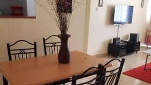 A dining area with a wooden table and chairs, a vase with branches, a flat-screen TV, and a red rug. - Stay.Plus Blue Bells Apartment Syokimau (Mlolongo)