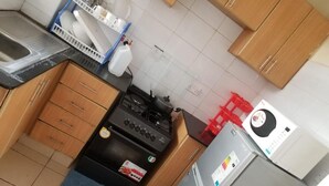 A compact kitchen with a gas stove, a built-in oven, a refrigerator, and wooden cabinets. - Stay.Plus Blue Bells Apartment Syokimau (Mlolongo)