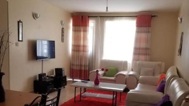 A living room with a TV, a dining table with chairs, a sofa, and a coffee table. - Stay.Plus Blue Bells Apartment Syokimau (Mlolongo)
