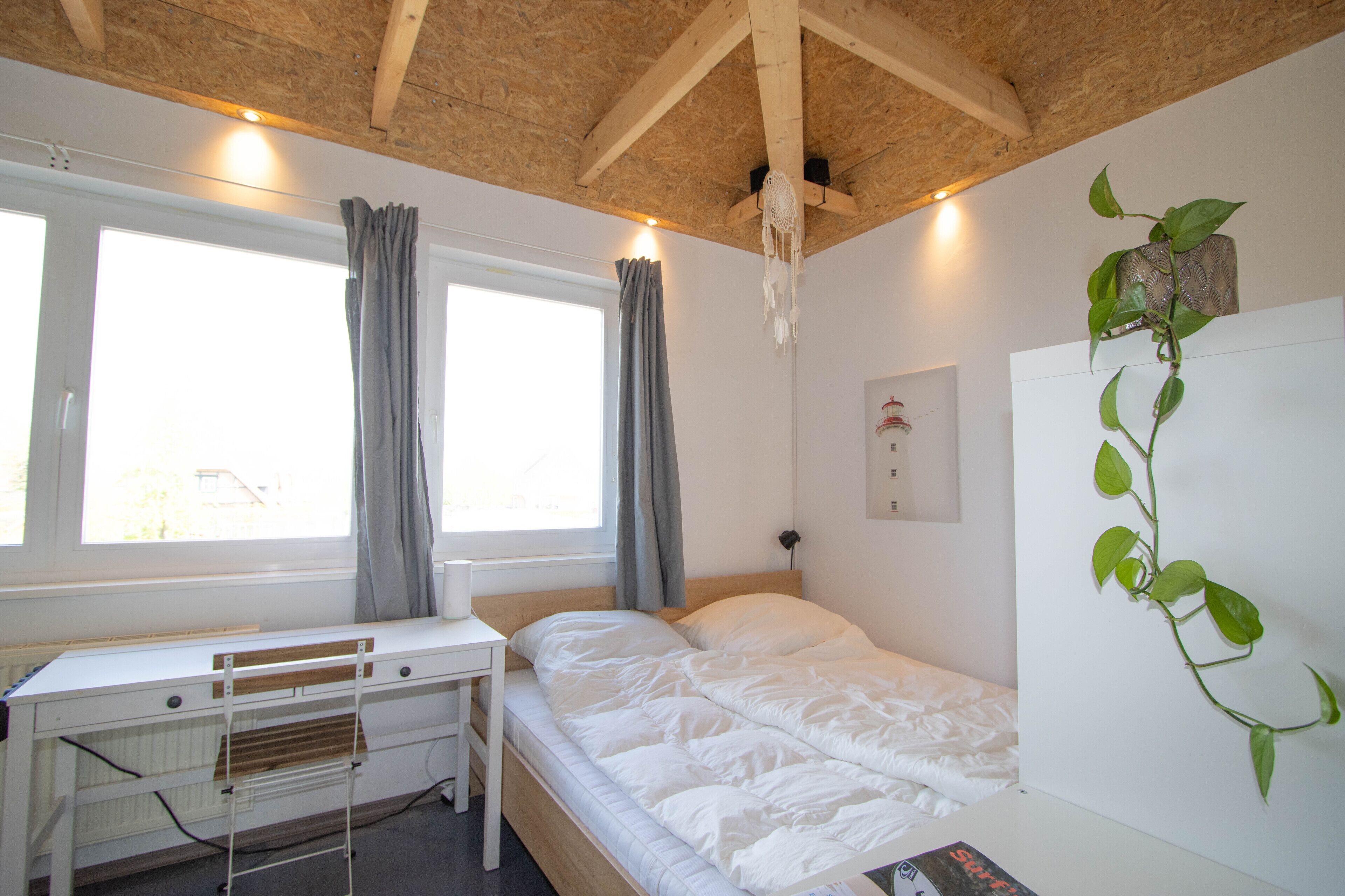A bedroom with a wooden ceiling, a white bed, a desk, and a window with curtains.