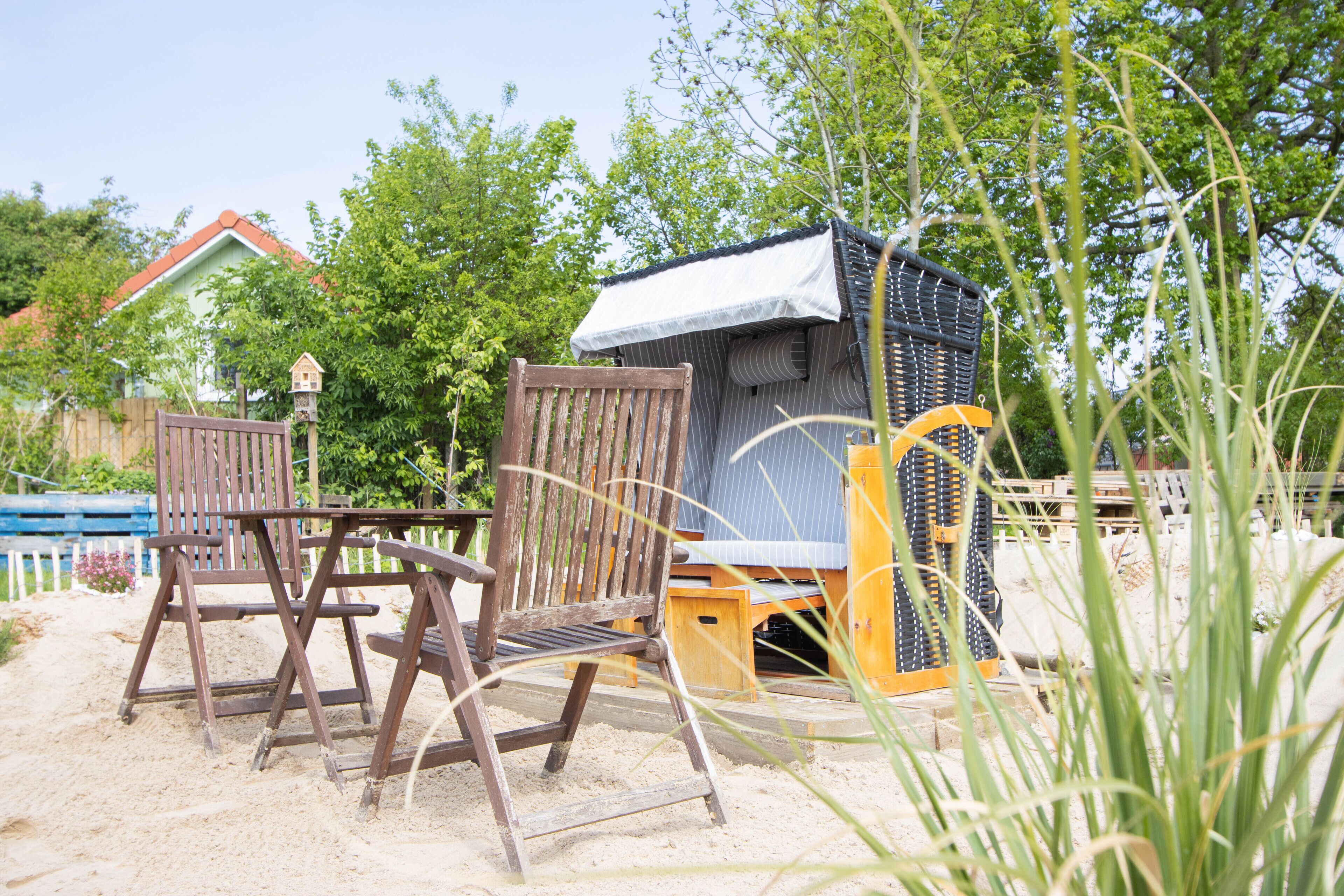A wooden chair and a small wooden structure in a sandy area with trees and a house in the background.