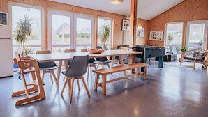 A spacious dining area with a long table, wooden chairs, and benches. There are large windows providing natural light and views of the outside.