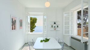 A bright, white dining area with a table, chairs, and a view of the sea through an open window.