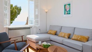 A modern living room with a grey sofa, wooden armchairs, a coffee table, and a view of the sea through an open window.