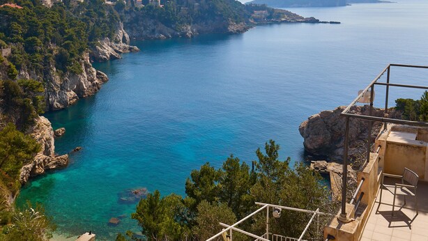 A coastal view with a clear blue sea, rocky cliffs, and a building with outdoor seating.