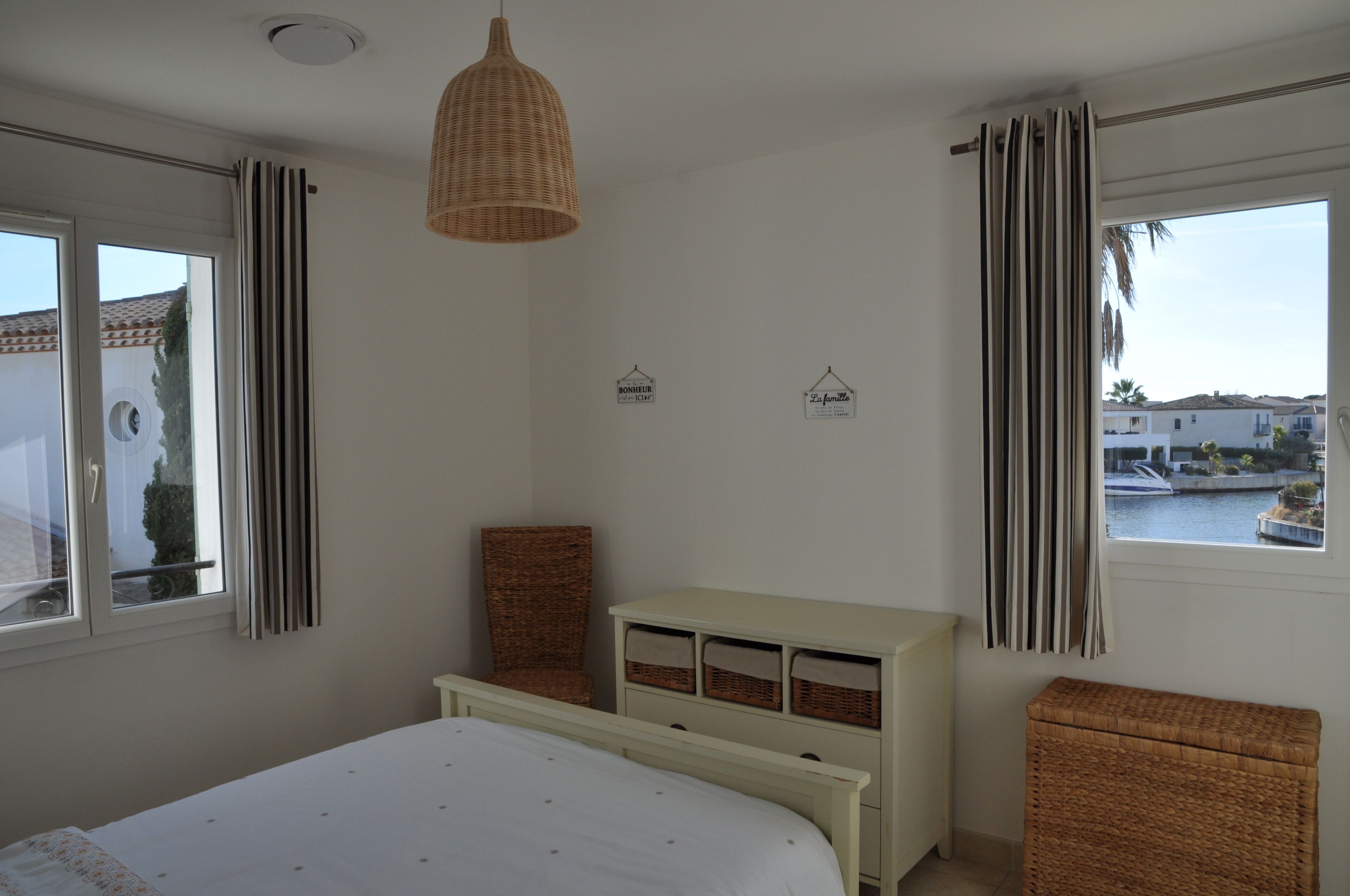 Une chambre à coucher avec un lit, une commode, un fauteuil en osier et une vue sur une étendue d’eau par la fenêtre.
