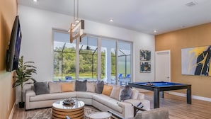A modern living room with a sectional sofa, a pool table, and a large window offering a view of the outdoors. - Premium Home w/ Hero Theme BR, Secret Gaming RM, Theater & Pool - Near Disney (Davenport)
