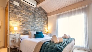 Ein Schlafzimmer mit Steinwand, einem Bett mit weißer Bettwäsche und türkisfarbenen Kissen, zwei Nachttischlampen und einem großen Fenster mit durchsichtigen Vorhängen.