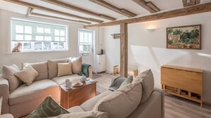 A cozy living room with a large beige sofa, a wooden coffee table, and a wooden cabinet. There is a window with a view of the outside, and a painting on the wall. - Candy Cottage, Halesworth (Halesworth)