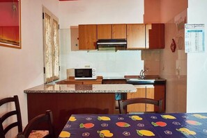 A compact kitchen with wooden cabinets, a microwave, and a sink. There is a dining table with a fruit-patterned tablecloth. - Cala Gonone, PT Casa i Pini, with giadino and parking.  (Cala Gonone)