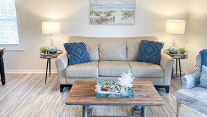 A living room with a beige sofa, two side tables with lamps, a coffee table with decorative items, and a beach-themed painting on the wall. - The Gathering Place - 1 Block from Beach! (Emerald Isle)