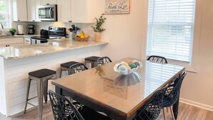 A modern kitchen with white cabinets, a granite countertop, and a dining area with a wooden table and black chairs. - The Gathering Place - 1 Block from Beach! (Emerald Isle)