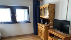 A room with wooden flooring, a television on a wooden stand, a cabinet with glass doors, and a window with curtains. - Studio apartment in front of Sassolungo mountain (Trentino-Alto Adige)
