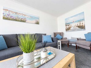 Sala de estar moderna com sofá cinzento, mesa de centro e duas poltronas. Há duas pinturas nas paredes, uma retratando uma paisagem costeira e a outra uma paisagem de praia.