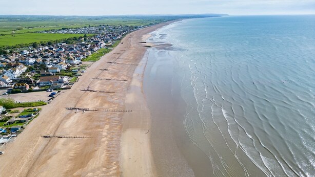 An aerial view of a coastal town with a long sandy beach, houses, and a clear view of the ocean.