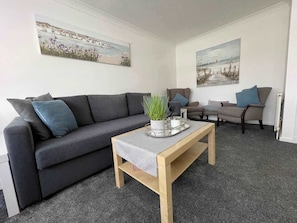 A living room with a grey sofa, a wooden coffee table, and two armchairs. There are two paintings on the walls.