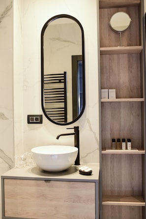 A bathroom with a white marble sink, a black faucet, a large oval mirror, and wooden shelves with toiletries.
