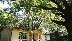 Une maison de plain-pied avec un porche, entourée d’une végétation luxuriante et de grands arbres.