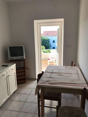 Et værelse med et spisebord, et fjernsyn på et stativ og udsigt til en bygning og grøntområder udenfor.