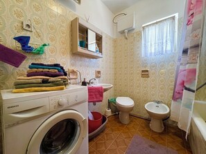Ein kleines Badezimmer mit Waschmaschine, Waschbecken, Toilette, Bidet und einem Fenster mit Vorhängen.