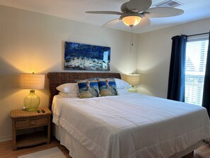 Une chambre à coucher comprenant un lit, deux tables de chevet, un ventilateur de plafond et une fenêtre avec des rideaux.
