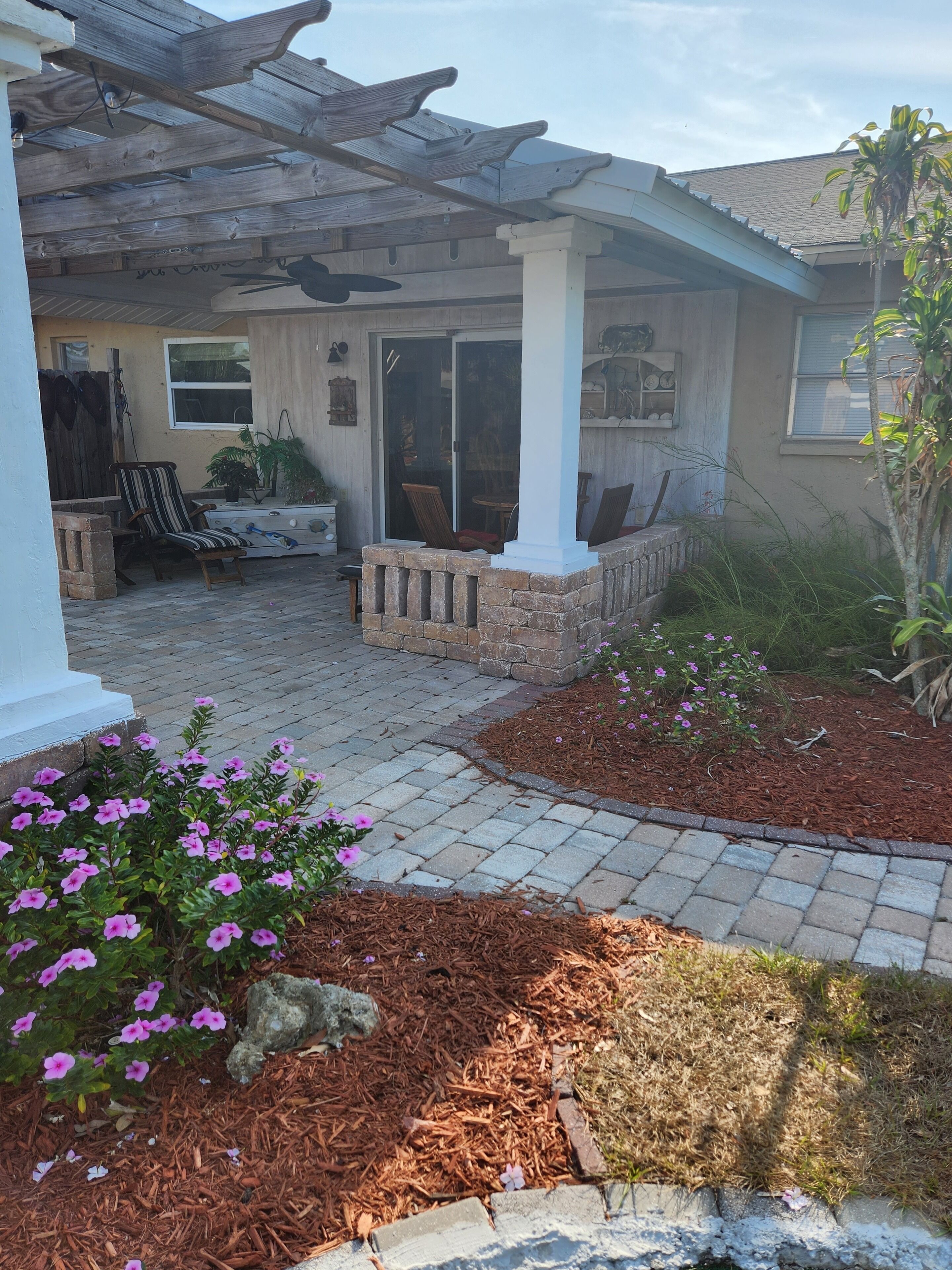 Ein überdachter Patio-Bereich mit Gartenmöbeln, einem Pflasterweg und blühenden Pflanzen.
