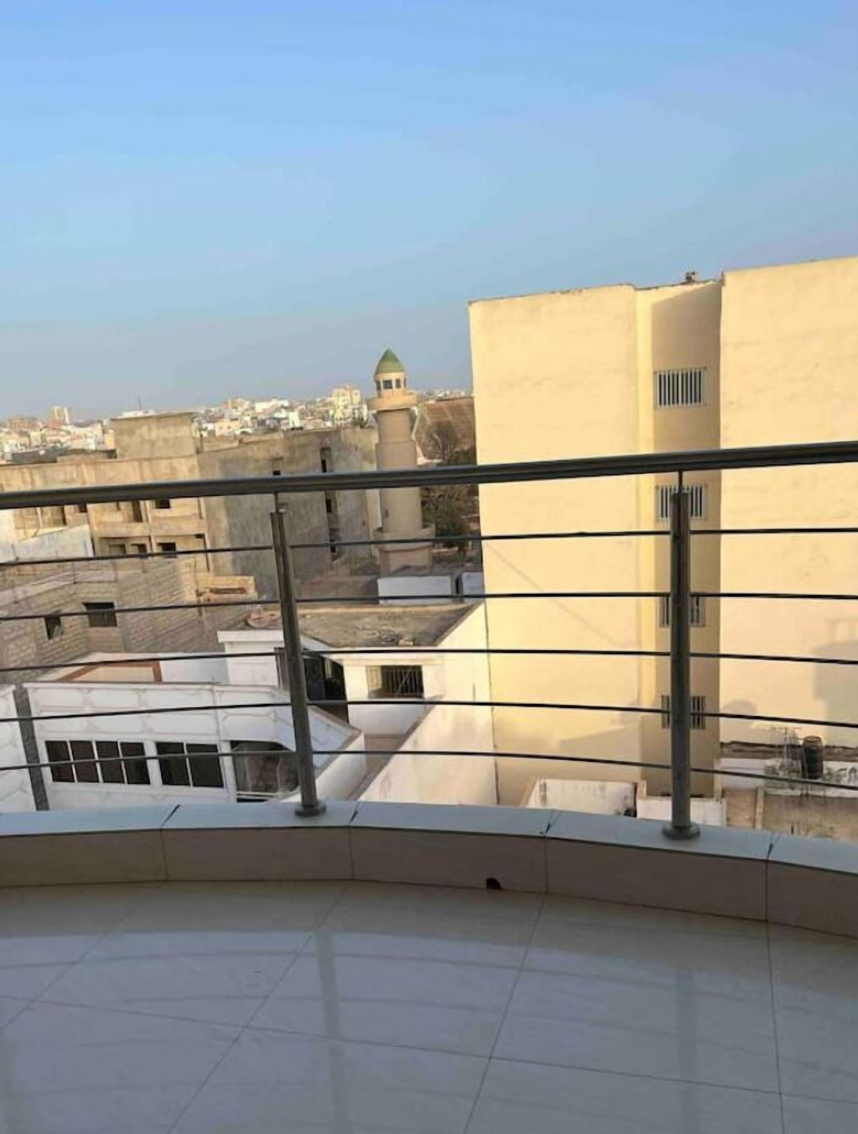 Vista desde un balcón con piso de baldosas, baranda de metal y un edificio con torre rematada en verde al fondo.