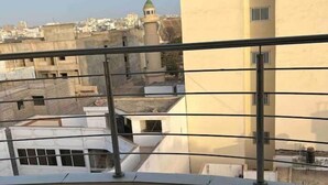 Une vue depuis un balcon doté d’un sol carrelé, d’une rambarde en métal, et d’un bâtiment surmonté d’une tour à toit vert, visible au loin.