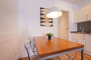 Un espace repas avec une table en bois, des chaises transparentes, une plante en pot, un luminaire suspendu et une cuisine en arrière-plan.