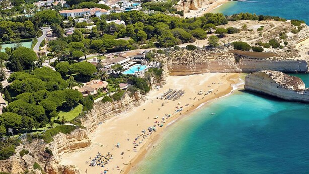 Uma área costeira com praia, falésias e zona de piscina rodeada por vegetação.