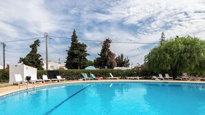 Piscina azul cristalina com cadeiras de lounge brancas e um pequeno edifício ao fundo.