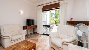 Uma sala de estar com dois sofás, uma mesa de centro de madeira, uma TV sobre um suporte de madeira, um ventilador e uma varanda com vista para vegetação.