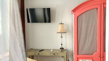 Een kamer met een houten plafond, een bureau met lamp, een stoel, een aan de muur bevestigde televisie en een kastje met bloemendecoratie.
