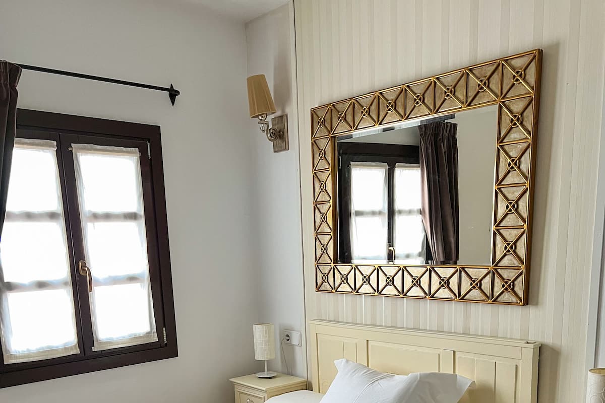 A neatly made bed with a patterned bedspread, a bedside table with a lamp, and a large mirror with a geometric frame.