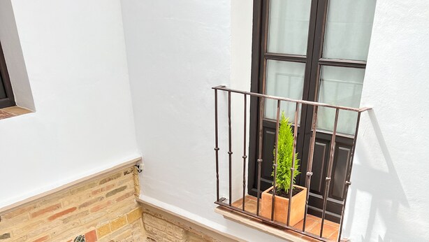 A courtyard with a brick floor, a small brick fireplace, a black door, and a window with a plant on the balcony. - Hotel Servilia (Carmona)