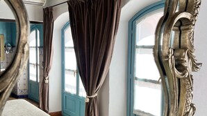 A room with a large ornate mirror, a bed with a patterned bedspread, and a view through a window with curtains. - Hotel Servilia (Carmona)