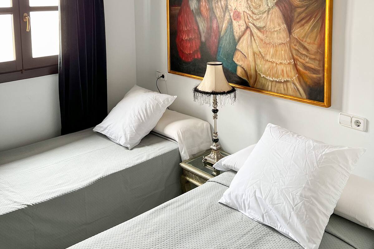A hotel room with two beds, a painting on the wall, a lamp, and a window with curtains.