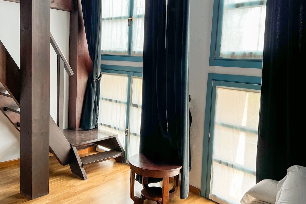 A room with a wooden staircase, a wooden chair, a bed, and large windows with curtains.