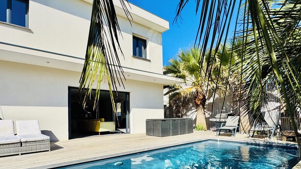 Un área de piscina moderna con una piscina azul cristalina, sillas de descanso y un edificio blanco con amplias ventanas.