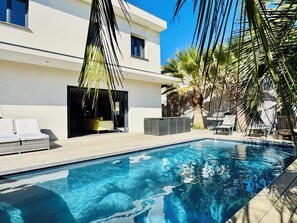 A modern pool area with a clear blue pool, lounge chairs, and a white building with large windows. - Modern Villa with Heated Pool – 50m from the Beach, Shops & Quiet Location (Agde)