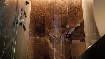 Un baño moderno con ducha envuelta en cristal, paredes de mármol y un tozaleto a la vista.