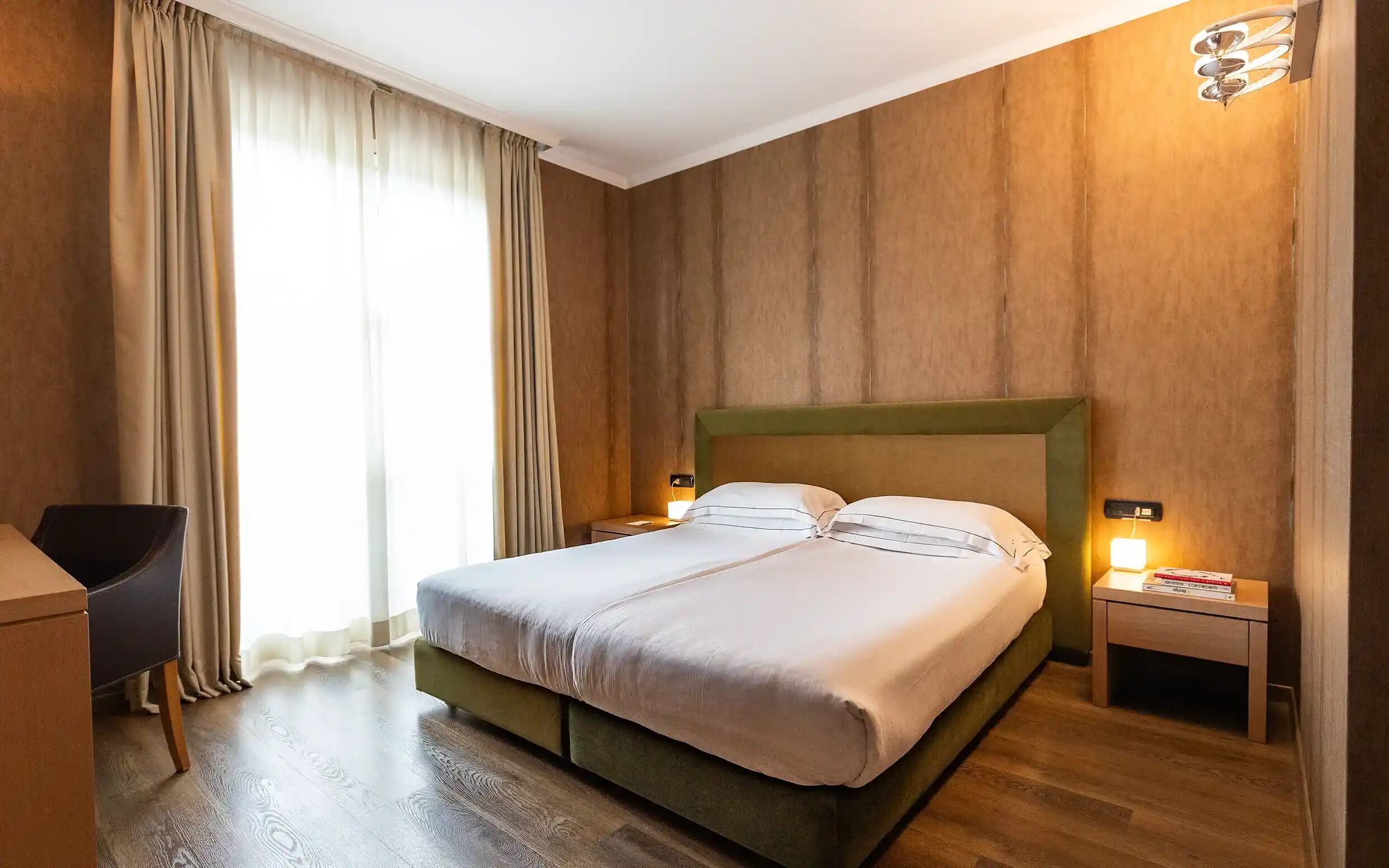 A hotel room with a double bed, wooden walls, a desk with a chair, and a nightstand with a lamp.