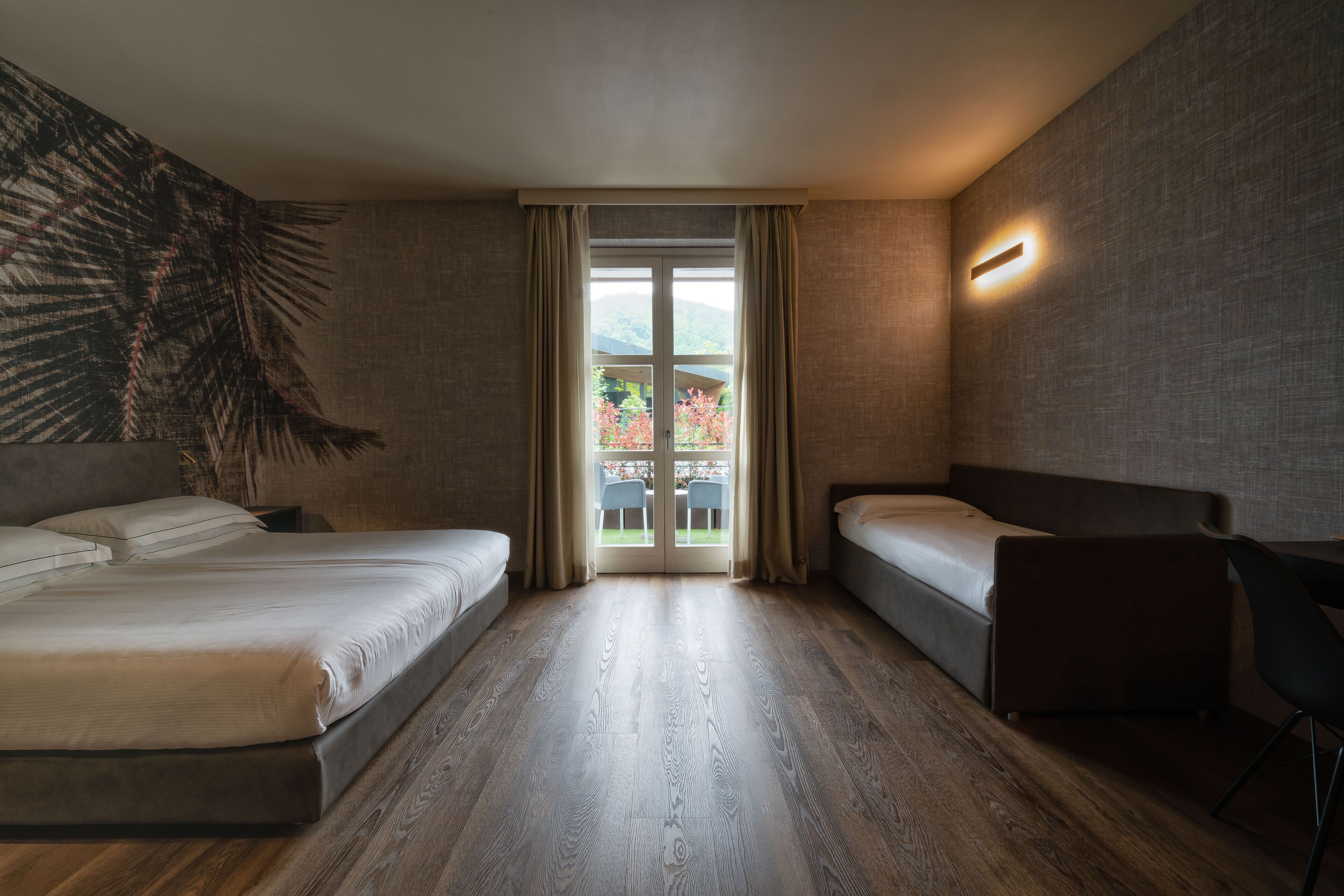 A modern hotel room with two beds, a wooden floor, a large window with curtains, and a wall-mounted light fixture.