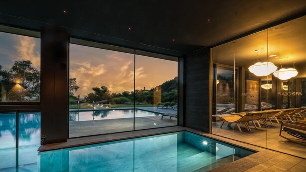 A modern indoor pool area with clear glass walls, lounge chairs, and a view of the sunset. - Phi Hotel Piajo (Nembro)